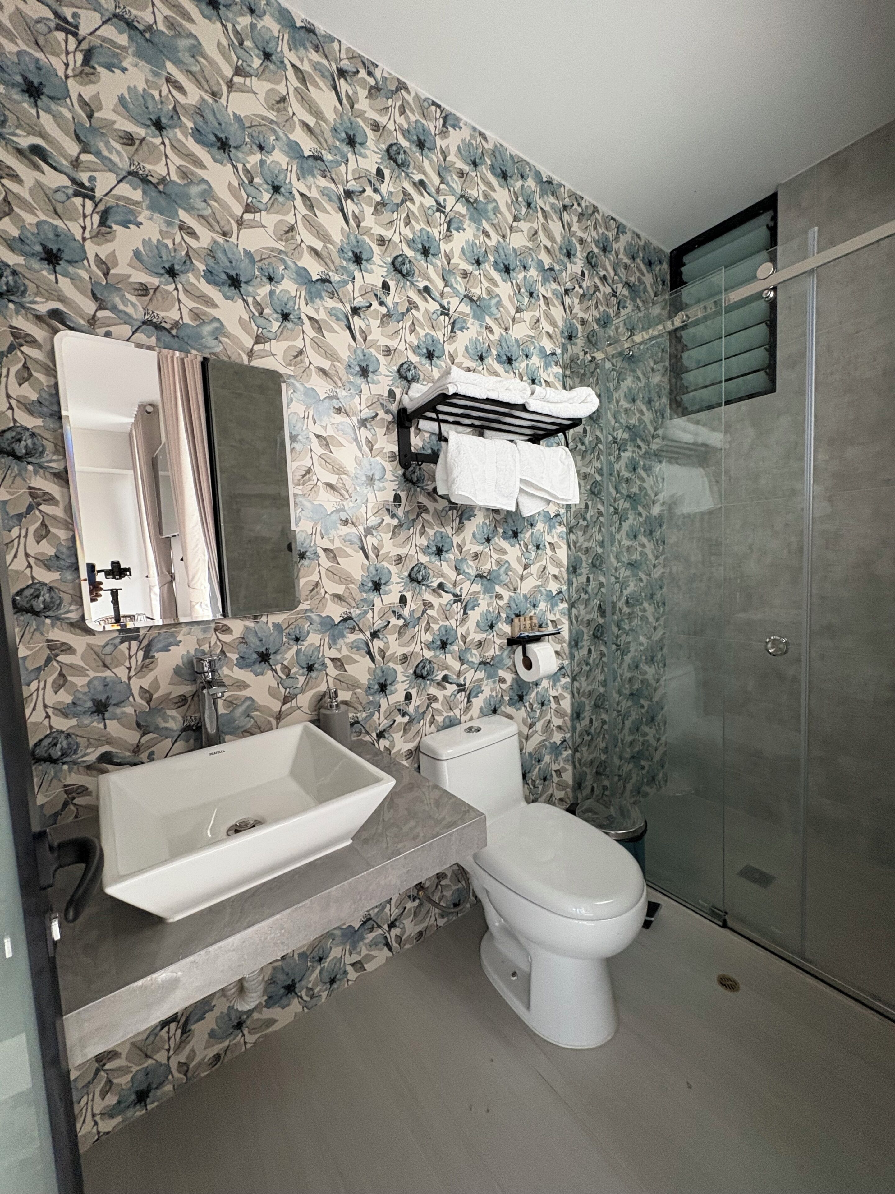 Une salle de bain avec un papier peint à motifs floraux, un lavabo blanc, des toilettes et une cabine de douche en verre.