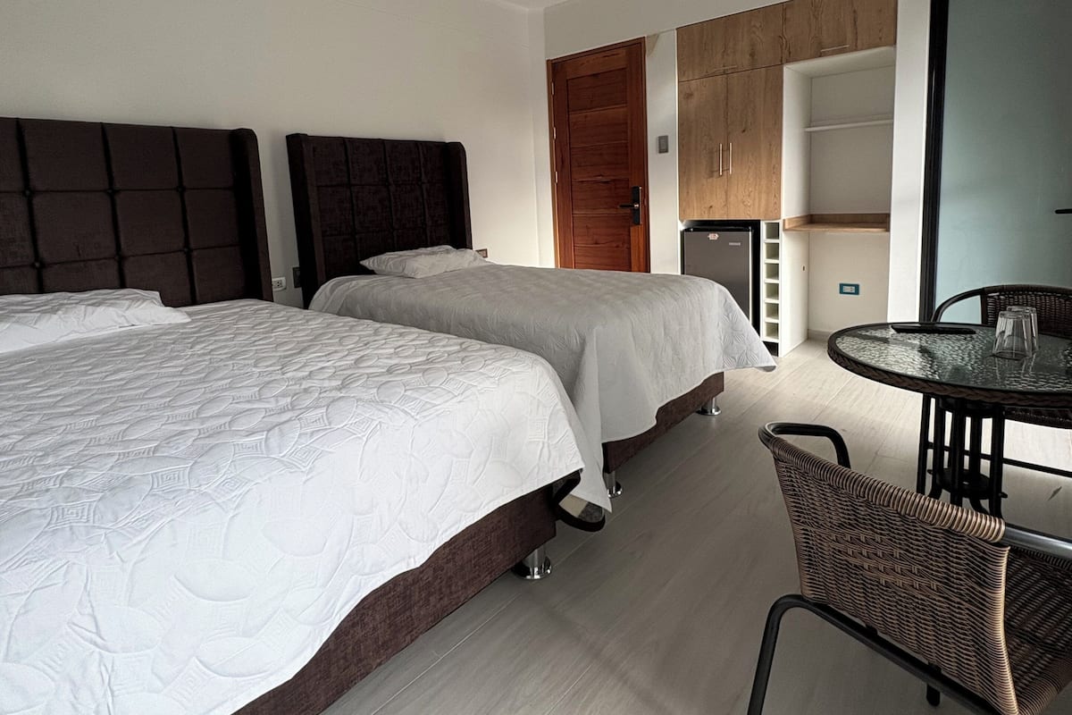 A modern hotel room with two beds, a dining area, and a kitchenette.