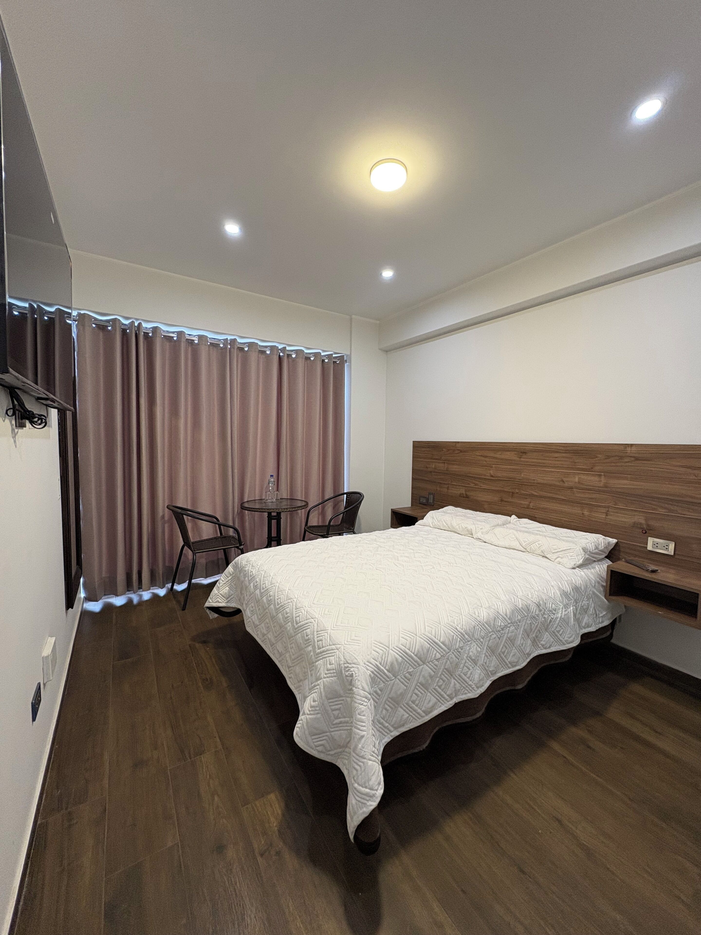 Un dormitorio con una cama, cabecera de madera, una mesita, dos sillas, un espejo y una ventana con cortinas.