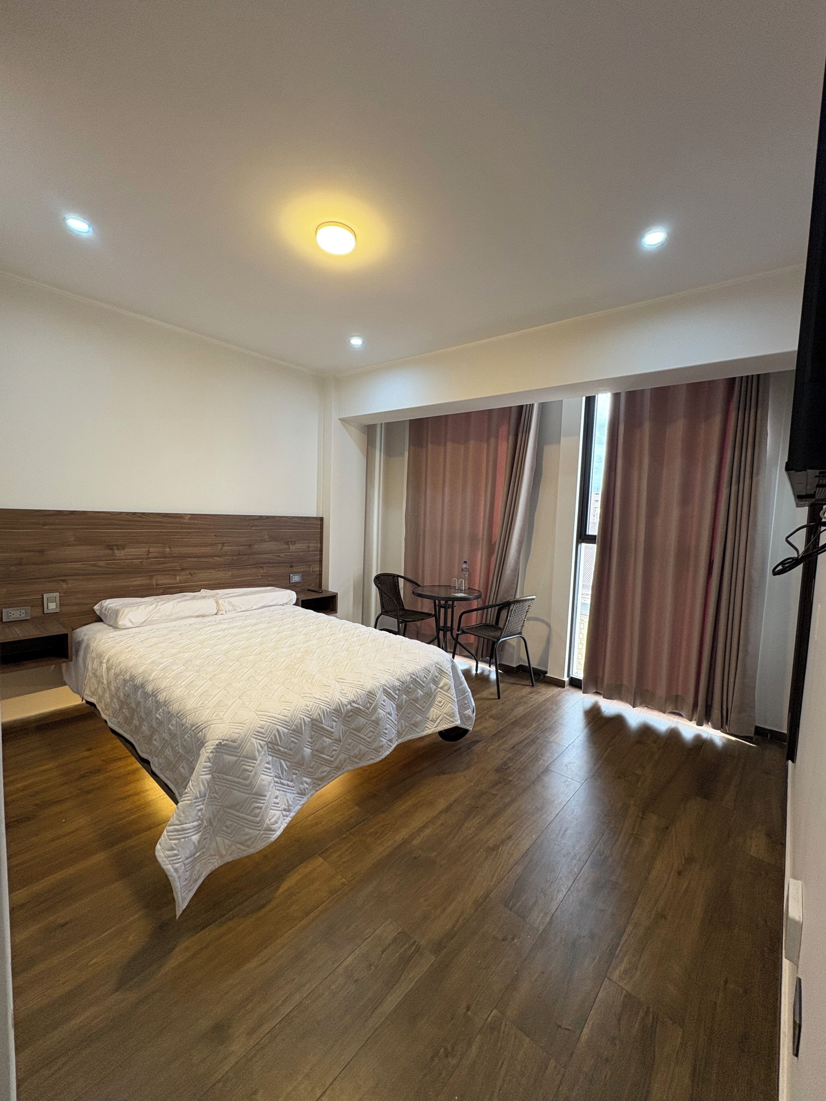 Habitación de hotel con una cama grande, una mesita con sillas, un televisor y una ventana con cortinas.