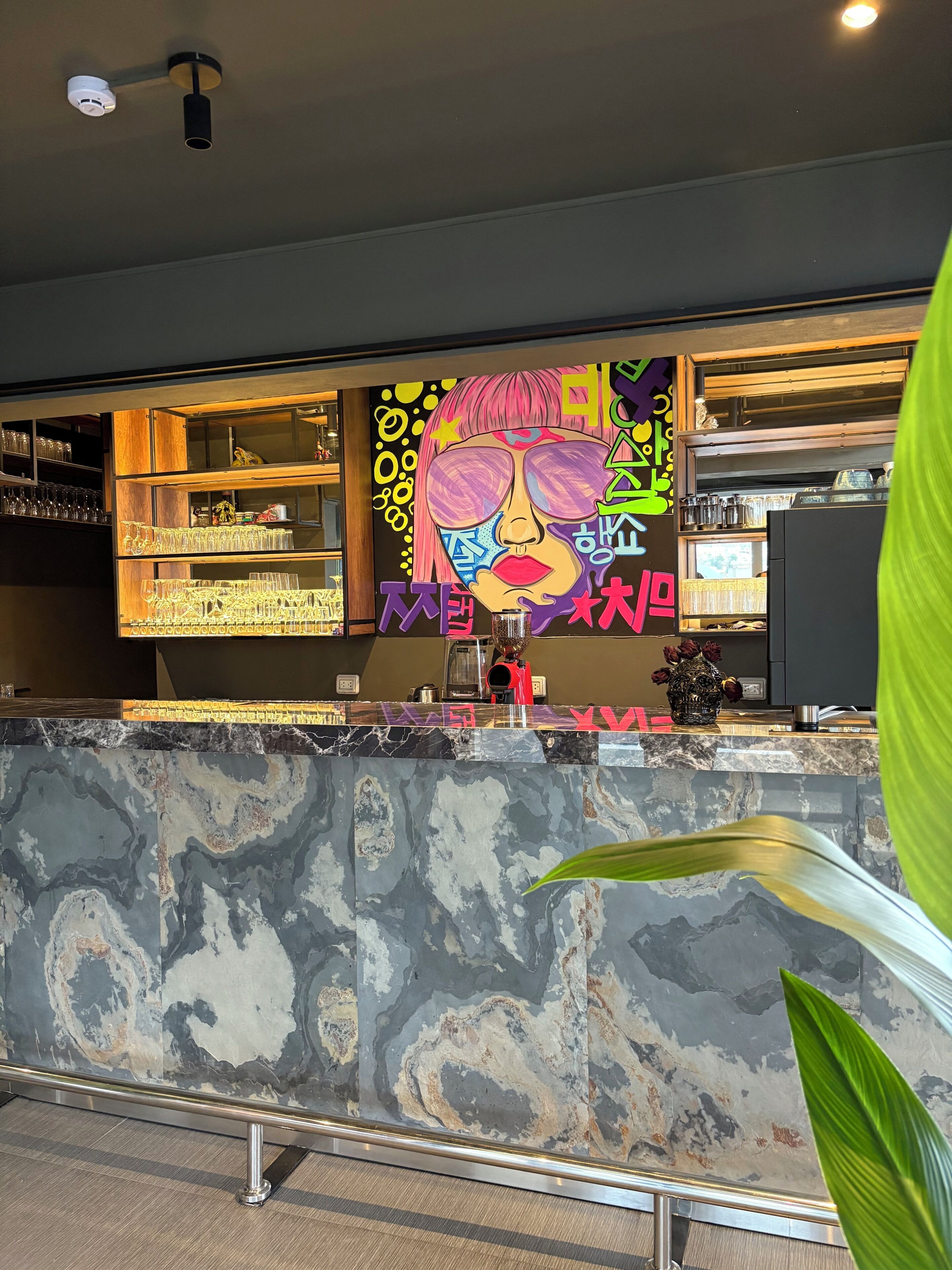 Una zona de bar con encimera de mármol, estanterías con copas y un mural colorido.