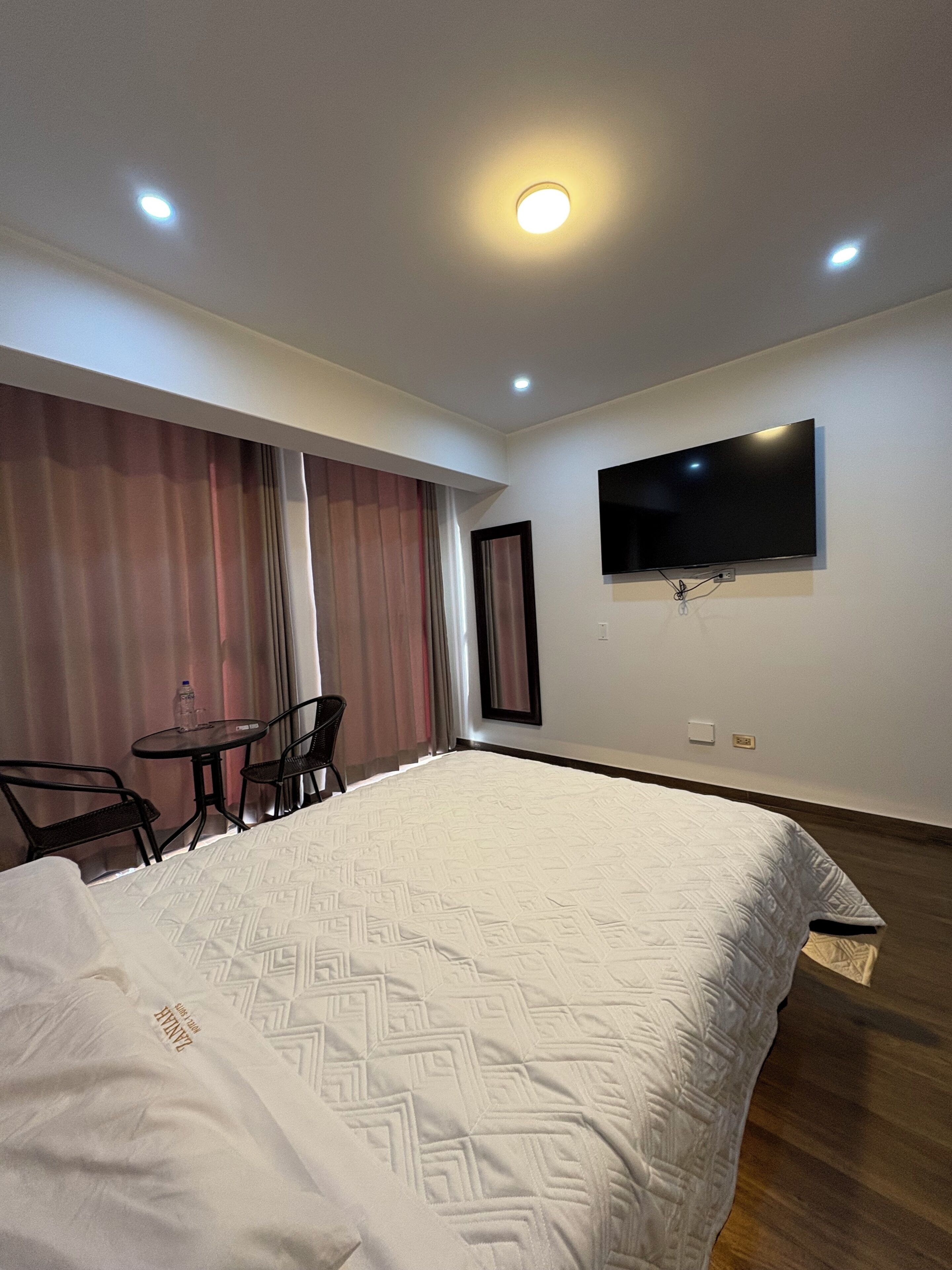 Ein Hotelzimmer mit einem großen Bett, einem an der Wand befestigten Fernseher, einem kleinen Tisch mit Stühlen und einem Fenster mit Vorhängen.