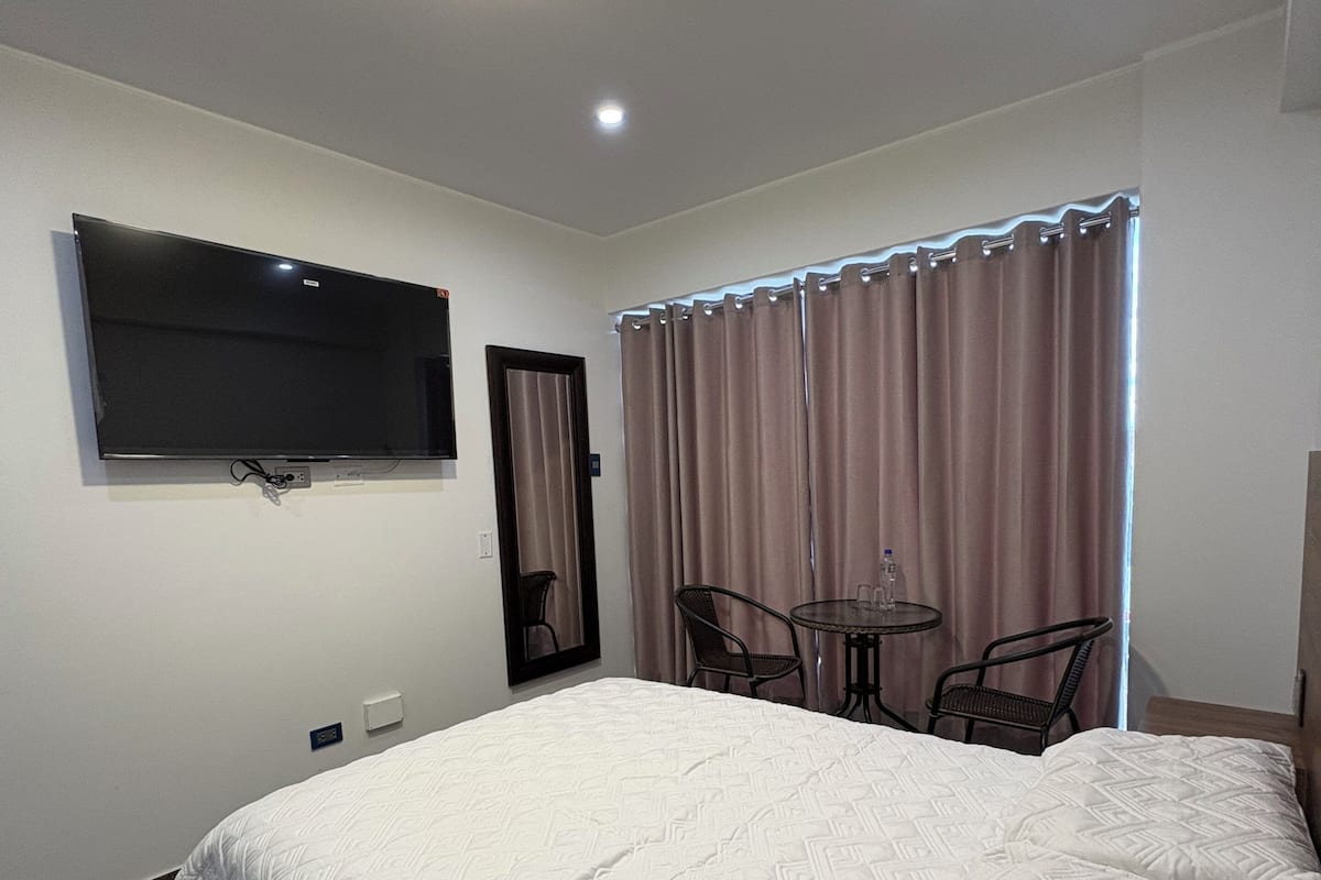 A hotel room with a large bed, a flat-screen TV mounted on the wall, a small round table with chairs, and a window with curtains.