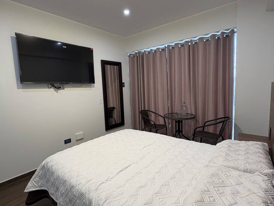A hotel room with a large bed, a flat-screen TV mounted on the wall, a small round table with chairs, and a window with curtains.