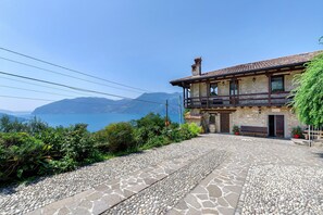 Une cour pavée de pierres, avec un bâtiment rustique doté d’un balcon et offrant une vue sur la mer et les montagnes.