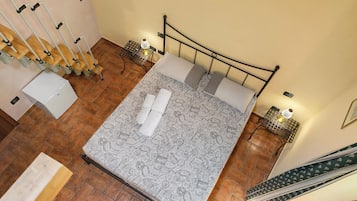 Ein ordentlich bezogenes Bett mit einem gemusterten Überwurf, zwei Kissen und zwei aufgerollten Handtüchern. Zwei Nachttische mit Lampen, ein Holzstuhl und ein kleiner Tisch mit Lampe sind ebenfalls vorhanden. Der Raum hat einen gefliesten Boden und eine hölzerne Sitzbank.
