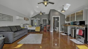 A spacious living area with a grey sofa, a coffee table, and a ceiling fan. There is a kitchen area with a stove, oven, and cabinets. - Quonset-Style Cabin & Sunset Patio Near Blue Hole (Wimberley)