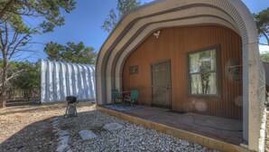 A rustic wooden building with a curved roof, a door, and a window. There is a small outdoor seating area with a table and chairs. - Quonset-Style Cabin & Sunset Patio Near Blue Hole (Wimberley)