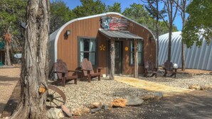 A rustic cabin with a curved wooden exterior, a large wheel, and outdoor seating. - Quonset-Style Cabin & Sunset Patio Near Blue Hole (Wimberley)