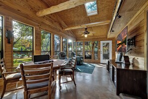Eine geräumige Holzhütte mit Essbereich, Deckenventilator, großen Fenstern und einem Fernseher.