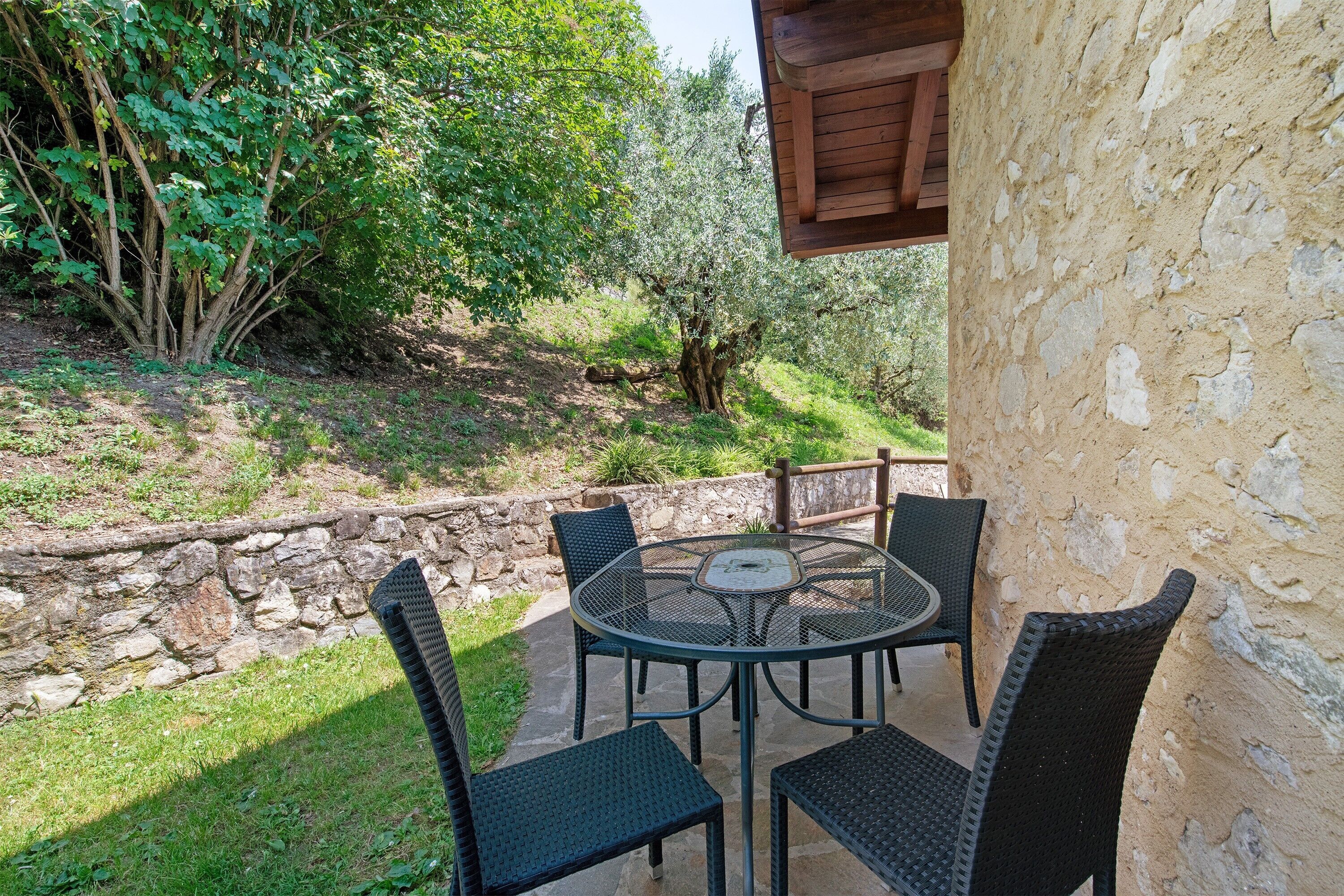 Eine Terrasse im Freien mit einem runden Tisch und Stühlen, umgeben von Steinmauern und viel Grün.