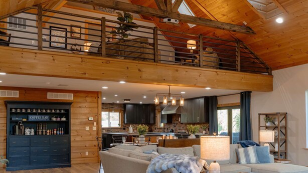 A spacious loft-style living area with a high ceiling and exposed wooden beams, a cozy seating area with a sofa and armchairs, a coffee bar with storage, and a kitchen in the background. - Compass Retreat for large groups (Buchanan)