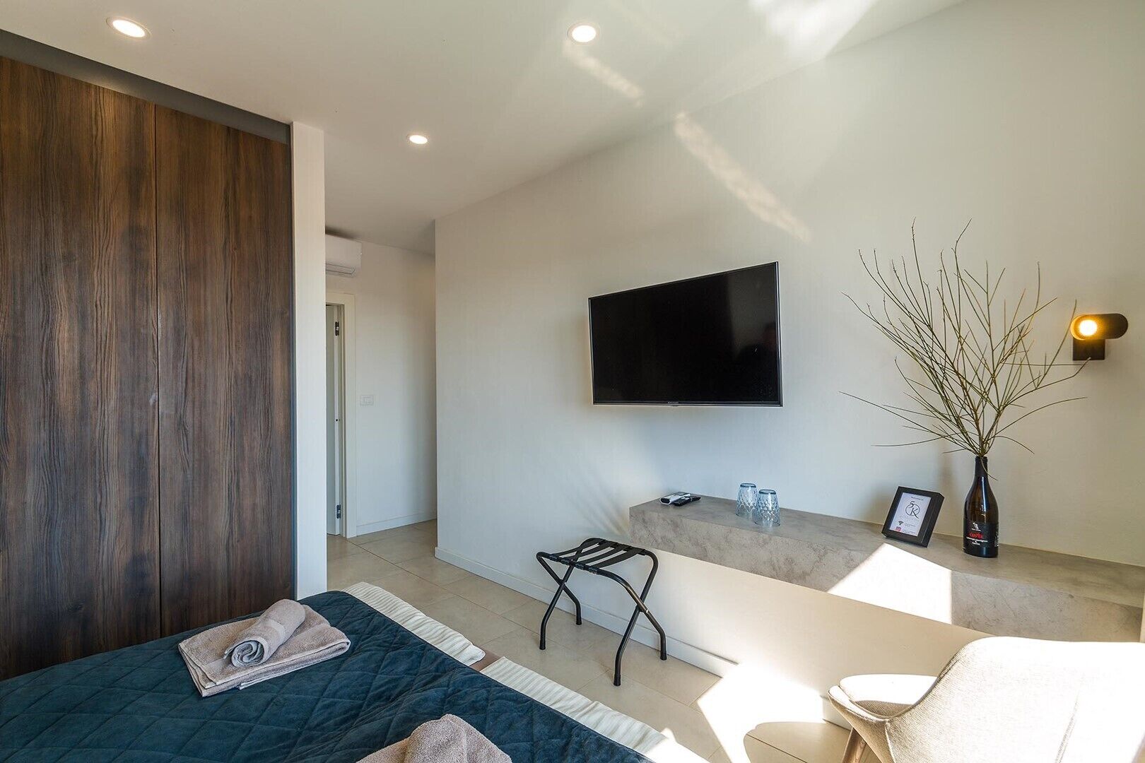 A modern hotel room with a bed, a small seating area, a TV, a bathroom vanity, and a plant.