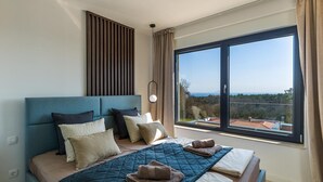 Een slaapkamer met een groot raam, een bed met veel kussens, een hoofdbord en een nachtkastje.