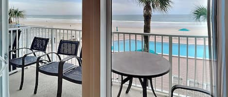 Un balcón con dos sillas de mimbre y una mesita, con vista a una playa y una piscina.