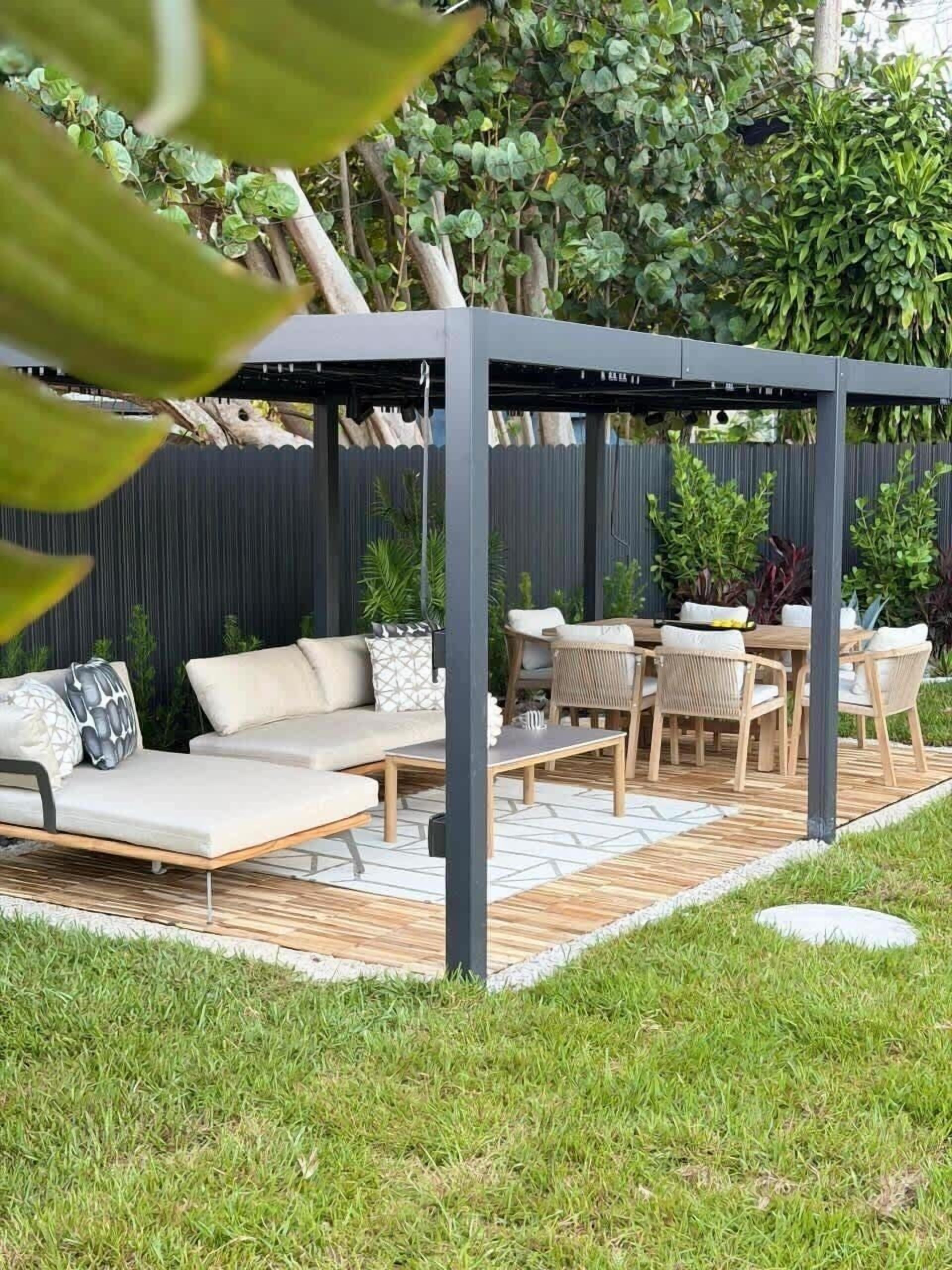 A covered outdoor seating area with a sofa, chairs, and a table, surrounded by greenery and a wooden deck.