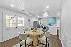 A dining area with a round table surrounded by chairs, a chandelier, and a view of the kitchen with blue cabinets. - Chic 2/2 Cottage Near Downtown & Beaches (Mount Pleasant)
