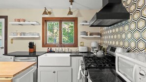 Una cocina moderna con encimera negra, armarios blancos, un lavamanos doble, una estufa y un microondas. Dos lámparas colgantes iluminan el lavamanos, y una ventana ofrece vistas a la vegetación.