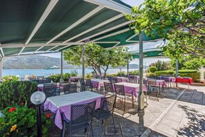 An outdoor dining area with tables and chairs, covered by a green canopy, overlooking a body of water and hills. - B&B Pension Kačina in Poljica - Mittel-Dalmatien / B&B Pension Kačina in Poljica - Doppelzimmer DD2 (Poljica)