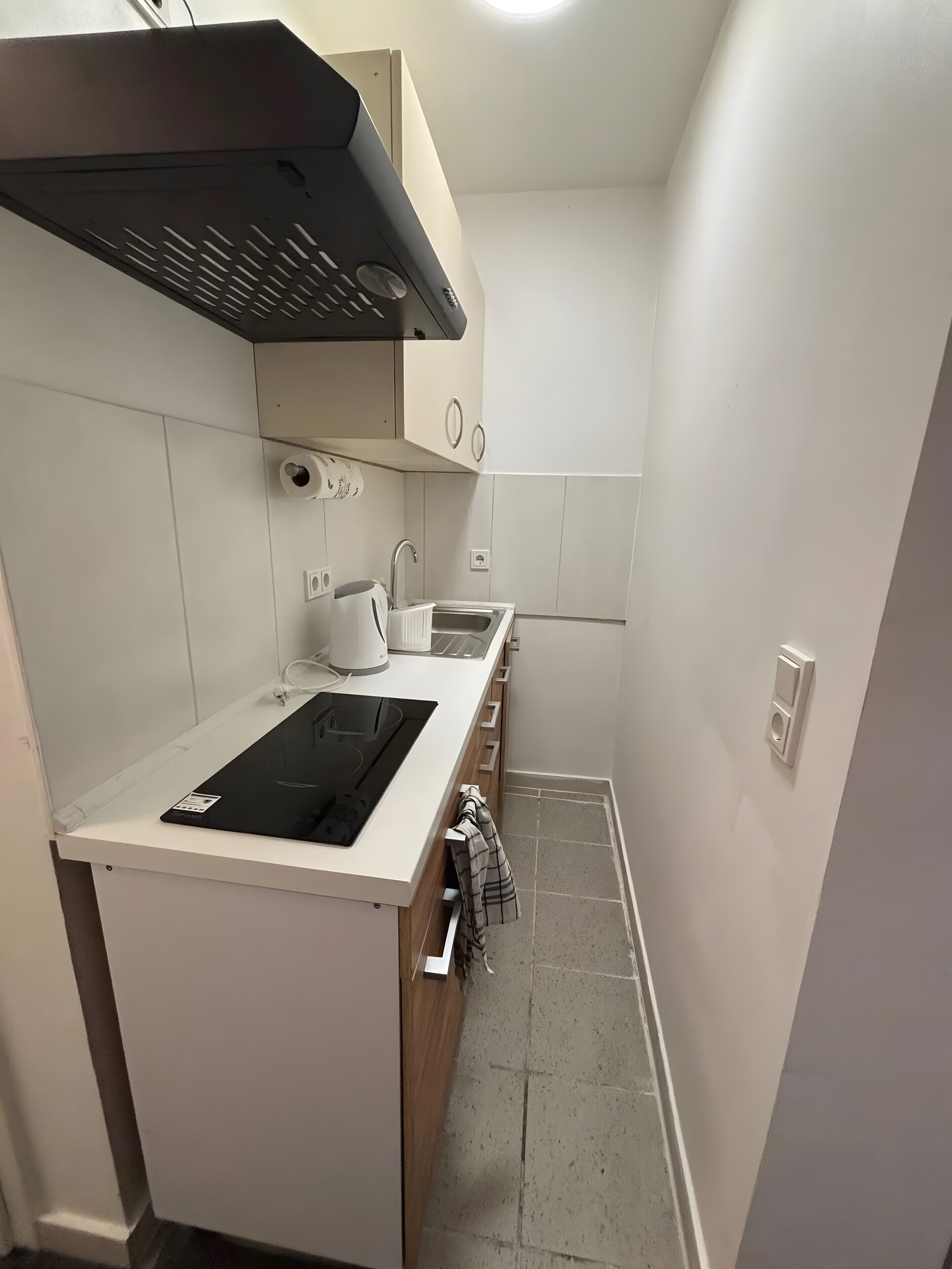 Una cocina compacta con cocina por inducción negra, encimera blanca, horno empotrado, pileta de acero inoxidable y extractor de aire negro.