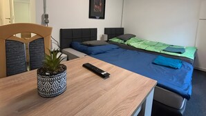 Una habitación pequeña y limpia con un escritorio de madera, una cama individual con ropa de cama azul, una planta en maceta y un cuadro en la pared.