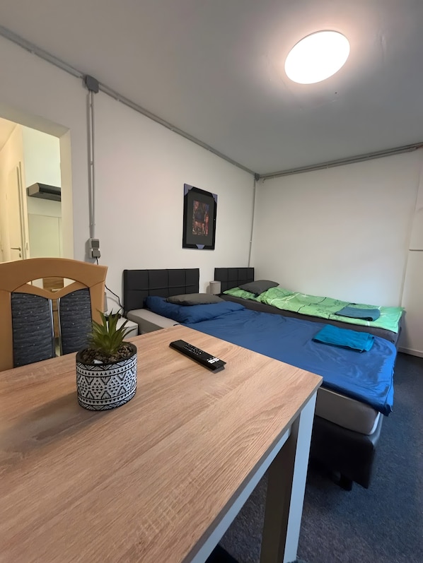 A small, clean room with a wooden desk, a single bed with blue bedding, a potted plant, and a framed picture on the wall.