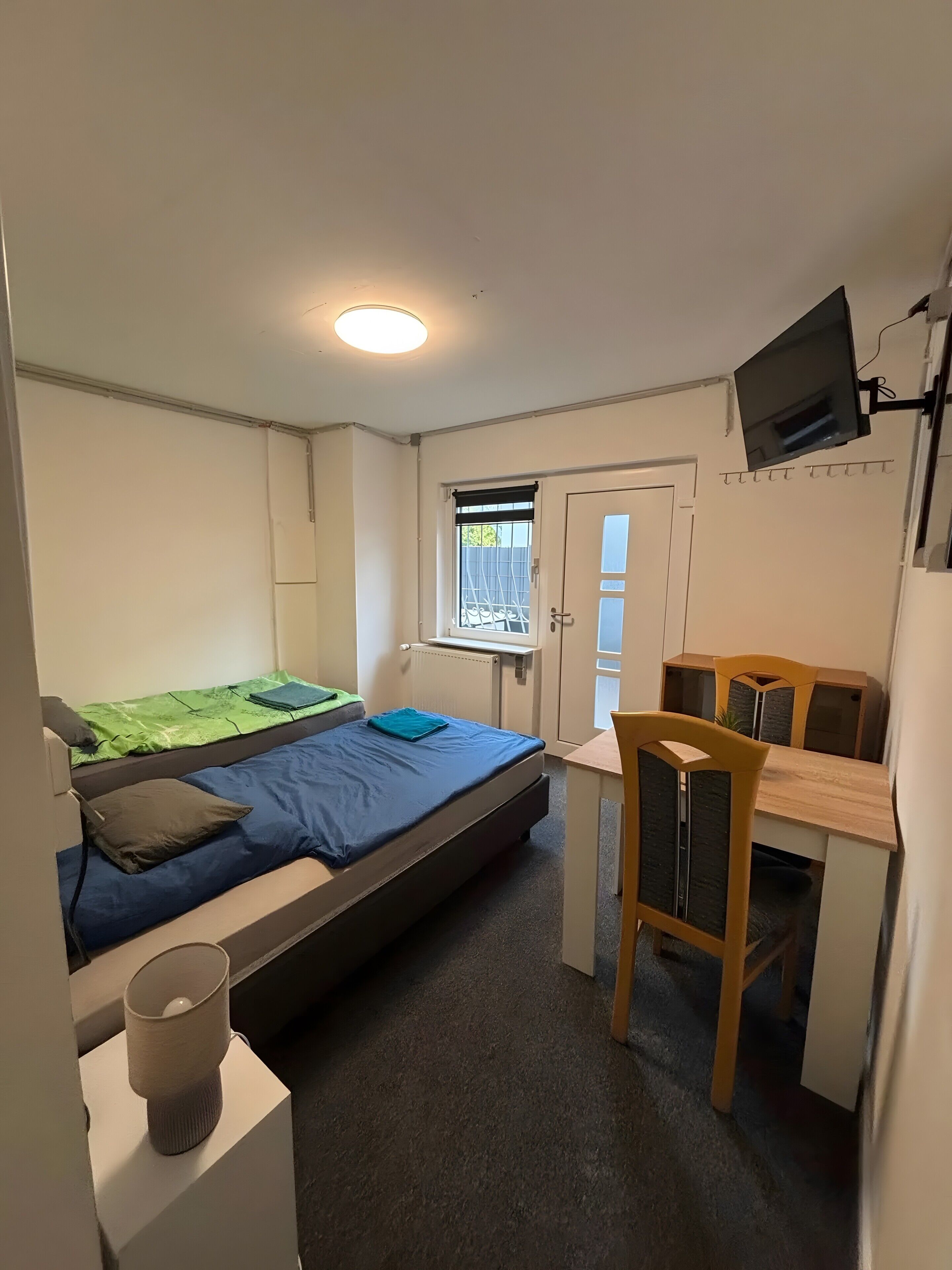 Une pièce de taille modeste comprenant un lit, un bureau, une chaise, un téléviseur fixé au mur et une fenêtre donnant sur un paysage agréable.
