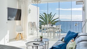 Una sala de estar moderna con balcón con vistas al mar, que incluye un sofá, una mesa de centro y un televisor montado en la pared.