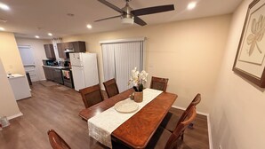 A dining area with a wooden table and chairs, a ceiling fan, and a kitchen in the background.