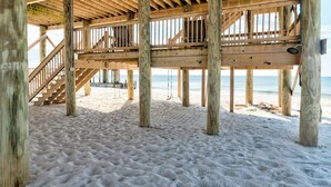 Una terraza de madera frente al mar con una escalera que sube a un nivel superior, sostenida por pilares de madera.