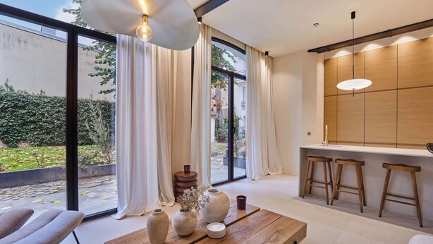 Ein modernes Interieur mit einem Holztisch, drei Hocker und großen Fenstern mit Vorhängen.