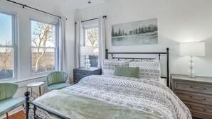 Un dormitorio con cama, mesita de noche, silla y ventana con cortinas.