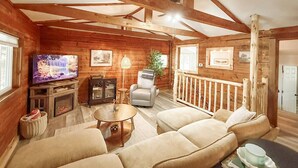 Een gezellig blokhutinterieur met een open haard, een televisie, een comfortabele bank en een houten balkenplafond.