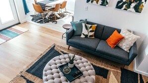 Un salon moderne avec un canapé gris, des coussins colorés, un pouf rond et un coin repas à l’arrière-plan.