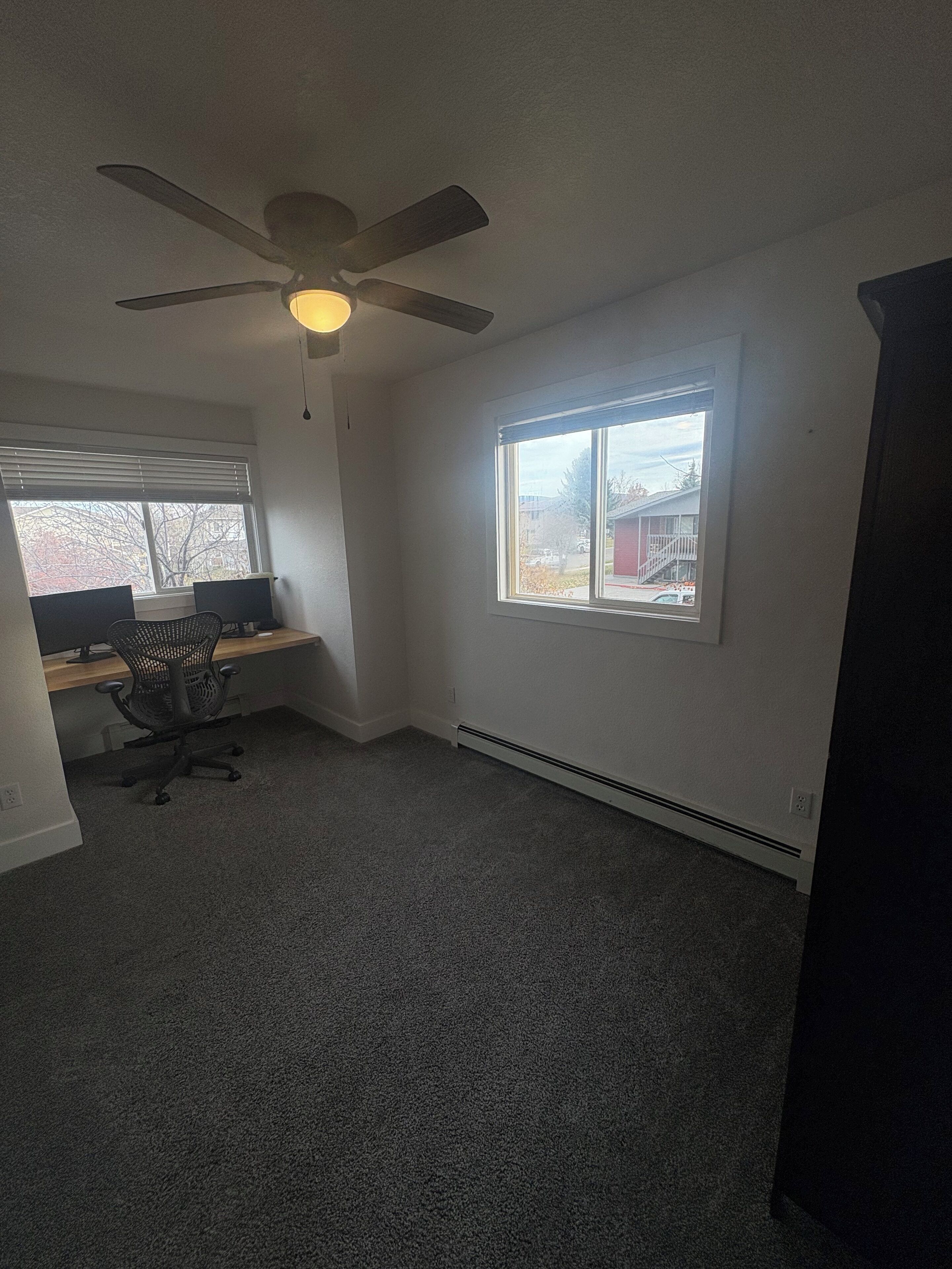 A room with a ceiling fan, a desk with two monitors, a black office chair, and a large window with a view of a building and trees.