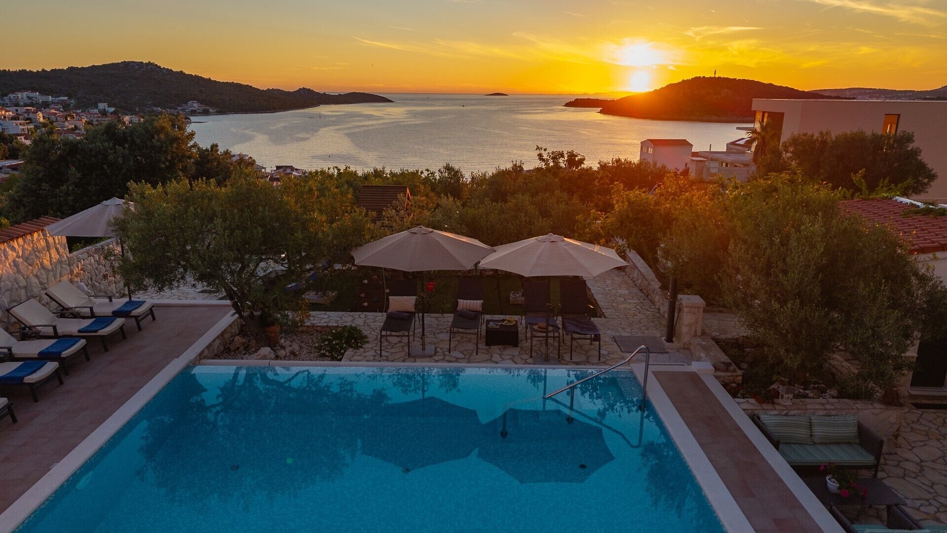 A swimming pool with lounge chairs and umbrellas, overlooking a body of water and hills during sunset.