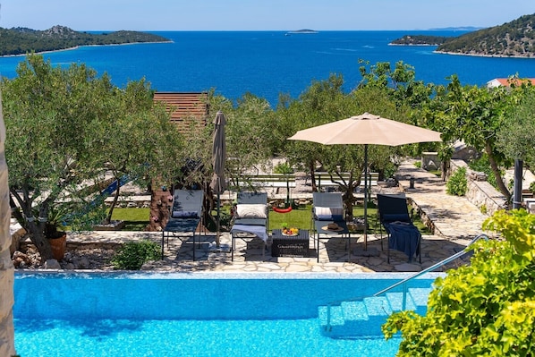Ein Außenpool mit Liegestühlen und Sonnenschirmen, umgeben von Bäumen und Blick aufs Meer.