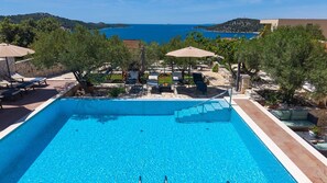 Ein rechteckiger Swimmingpool mit klarem, blauem Wasser, umgeben von Liegestühlen und Sonnenschirmen, mit Blick auf das Meer und die Hügel im Hintergrund.