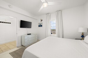 Ein Schlafzimmer mit einem Bett, einem Fernseher, einem Schminktisch, einem Fenster mit Vorhängen und einem Deckenventilator.