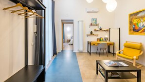 Un pasillo que conduce a una habitación con un escritorio, una silla y un sillón amarillo. Se observan prendas colgadas en un perchero y un cuadro en la pared.