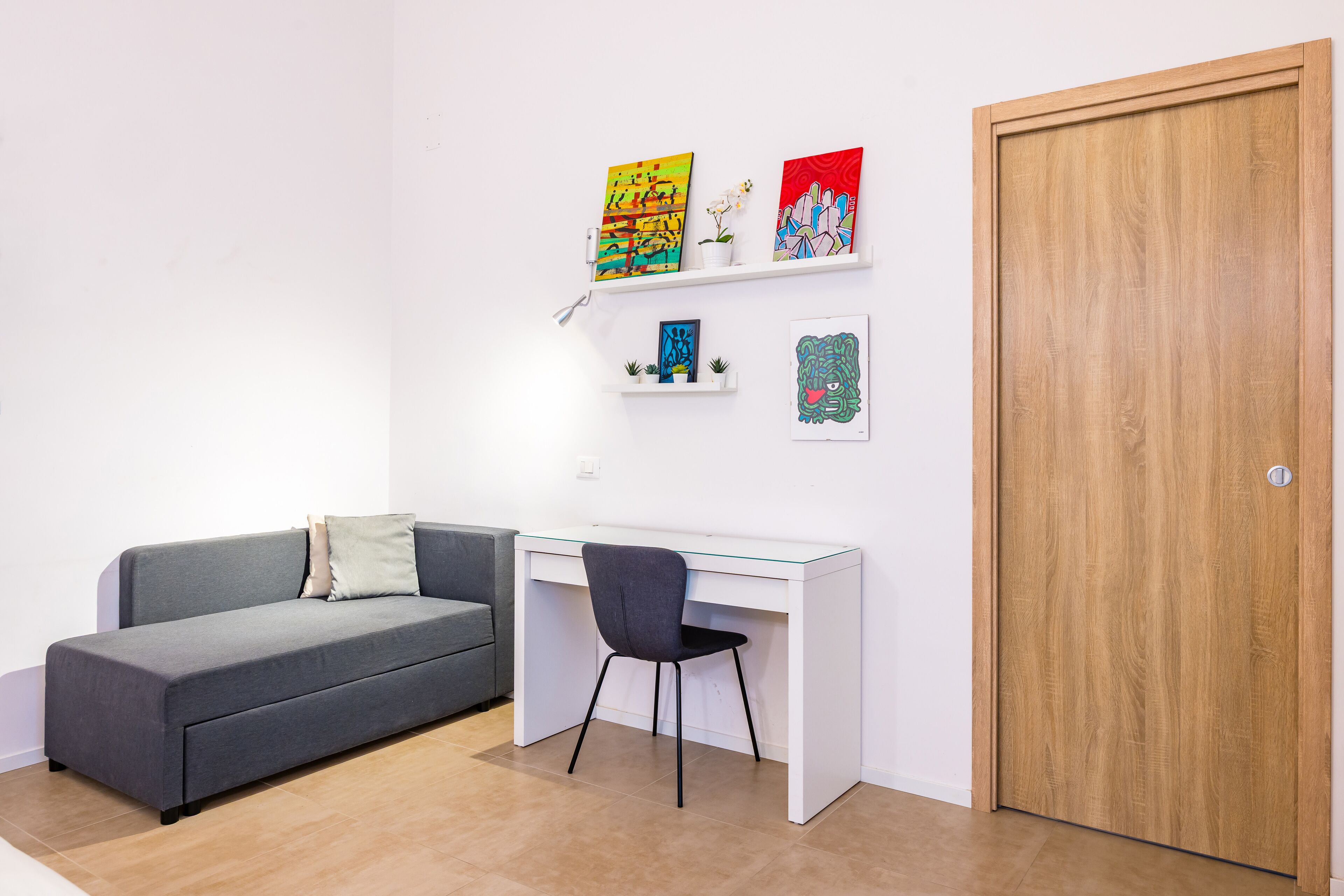 A modern room with a grey sofa, a white desk, and a wooden door. There are two shelves with artwork and a small plant.
