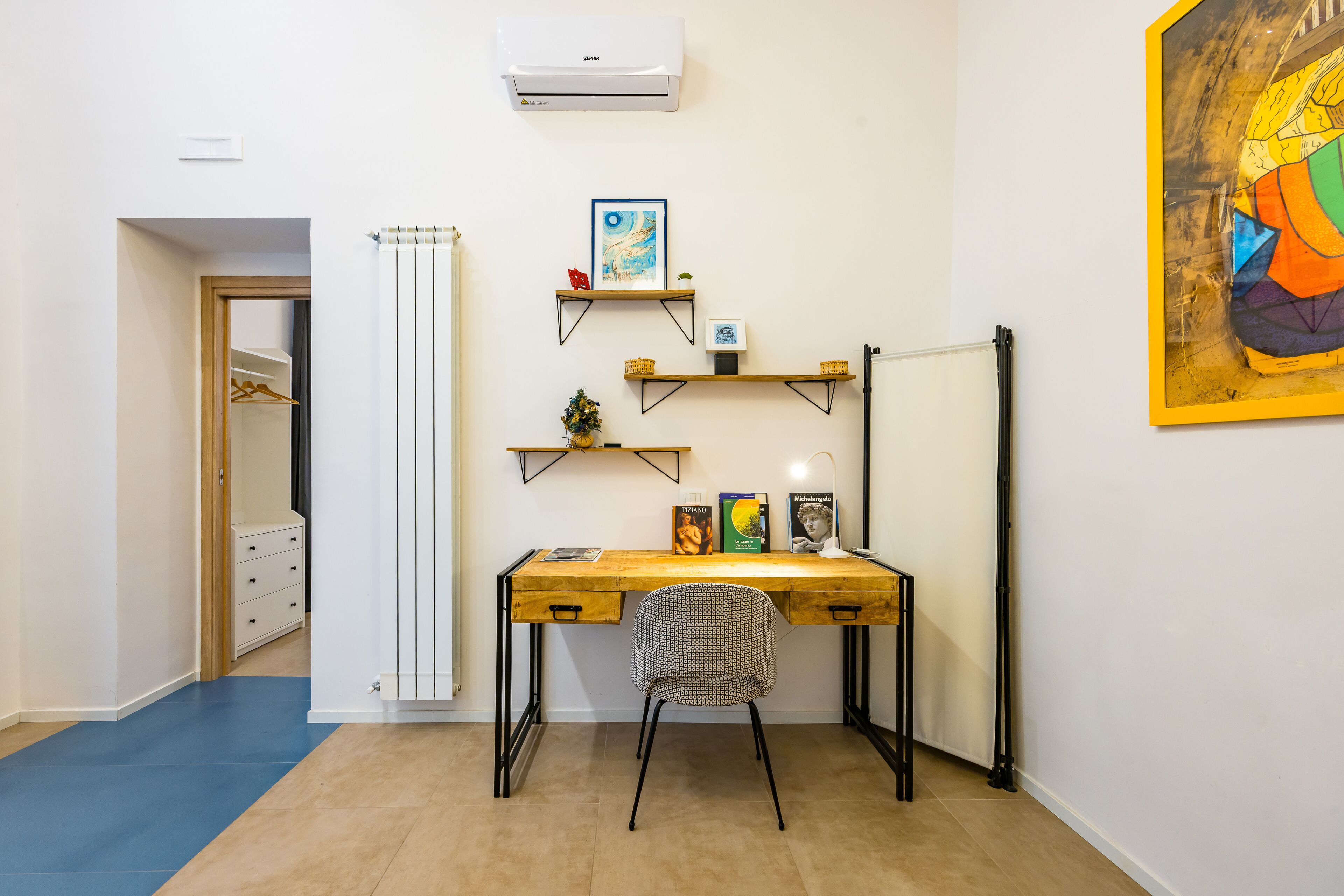 A compact room with a wooden desk, a chair, and a radiator. Shelves with books and decorative items are mounted on the wall.