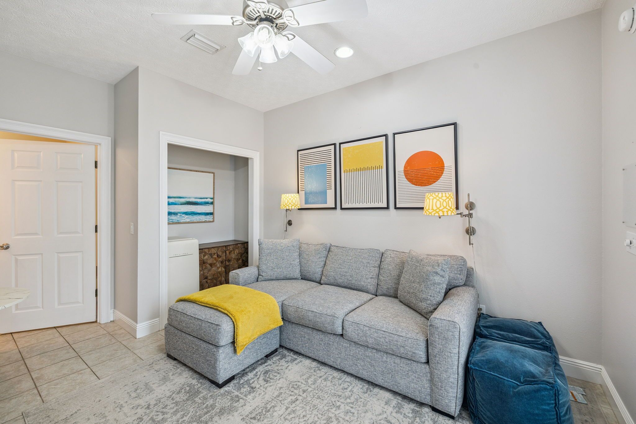 Un salon moderne avec un canapé d’angle gris, un plaid jaune et un pouf bleu. Des œuvres d’art encadrées ornent le mur, et un ventilateur de plafond est installé au-dessus.