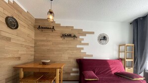 A cozy room with a wooden wall, a wooden table with chairs, a red sofa, and a small shelf. - Bright T2 with mountain view and comfort (La Salle-les-Alpes)