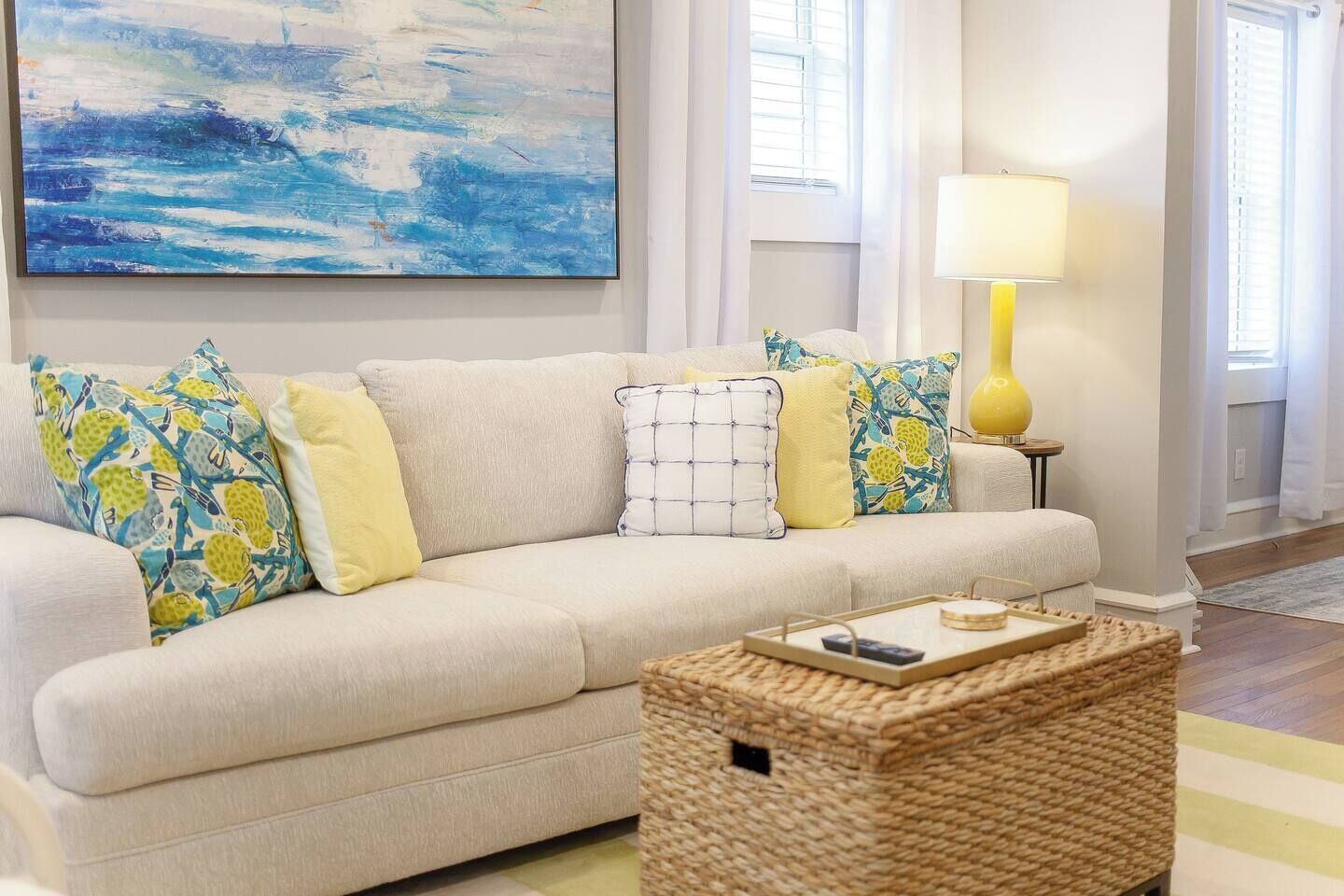 A living room with a beige sofa, patterned pillows, a painting, a lamp, and a woven basket.