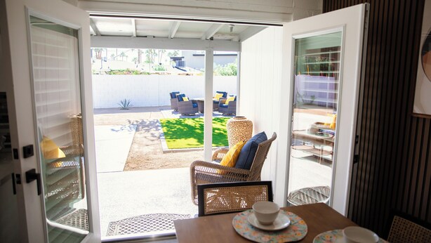 Une porte coulissante ouverte donne sur un patio aménagé avec des meubles en osier et une table préparée pour deux personnes.