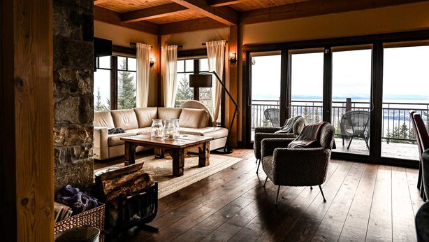 A cozy room with a stone wall, wooden ceiling, large windows, a sofa, a table, and a chair. - Magique | Hot Tub & 360° St. Lawrence River View (Les Éboulements)