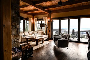 A cozy room with a stone wall, wooden ceiling, large windows, a sofa, a table, and a chair. - Magique | Hot Tub & 360° St. Lawrence River View (Les Éboulements)