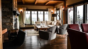 A cozy living room with a stone fireplace, wooden beams, a sofa, and a view of trees through large windows. - Magique | Hot Tub & 360° St. Lawrence River View (Les Éboulements)
