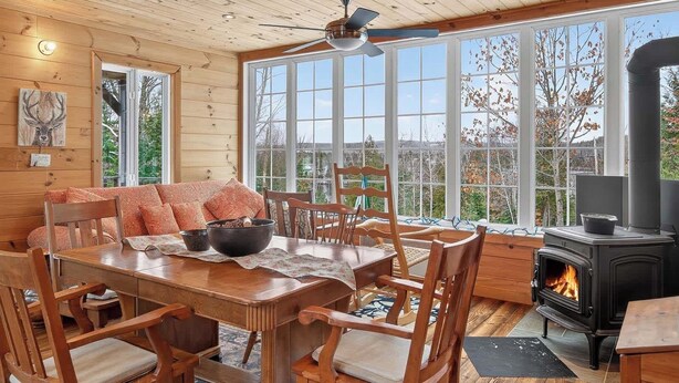 A cozy cabin interior with a dining area, wooden furniture, a fireplace, and large windows offering a view of the outdoors. - Nature & Comfort Chalet on Lake Chapleau (La Minerve)